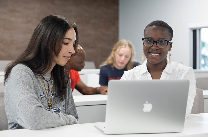 Two people sitting inside a room and looking at an Apple laptop